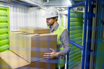 Young man in work helmet taking cardboard box and loading it into the chamber in warehouse