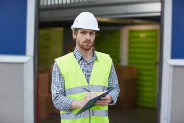 Portrait of young worker in work helmet and in reflective vest looking at camera while making notes and making delivery outdoors