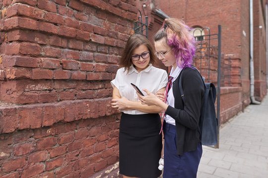 Female student walking and talking with woman teacher, girl showing on smartphone screen