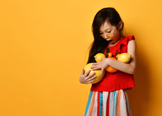 Little asian girl in sunglasses, brown beret, red blouse, colored skirt. Excited, holding pomelo, oranges and lemons. Studio shot.