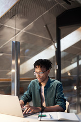 African young man in eyeglasses sitting at the table and busy with his work he typing on laptop computer at modern office