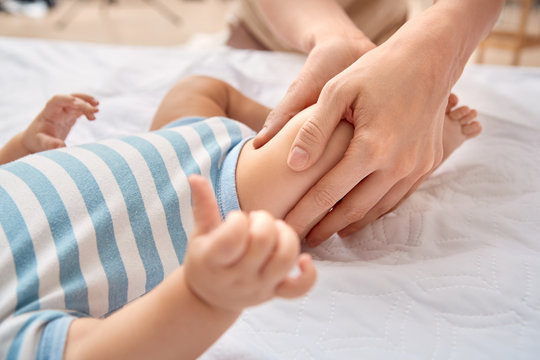 Baby Care. Young mother doing pediatric massage massaging legs of little son lying on bed close-up