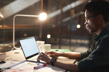 Serious African man sitting at the table with documents and typing on laptop computer till late at...