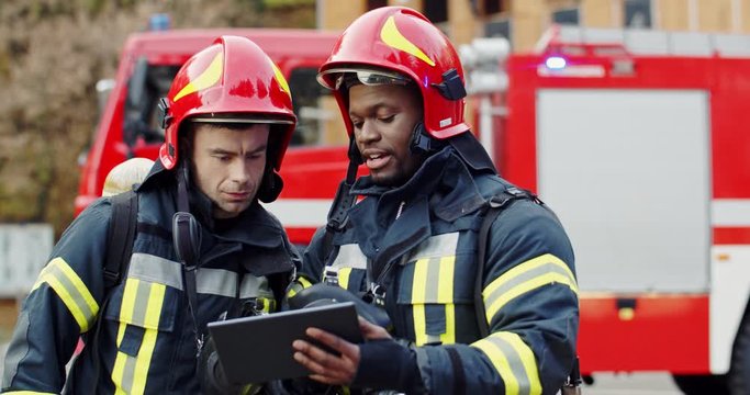 Caucasian and African American firemen in full equipped clothes standing outside at the fire truck with a plan in hands and deciding what to do for extinguishing fire.