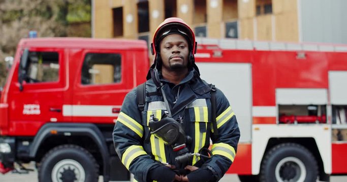 Portrait shot of the young African American handsome fireman in costume and helmet standing on the background with fire truck. Outdoors.