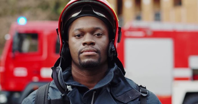 Close up of the serious face of teh young African American man fire fighter in helmet looking straight to the camera. Outside. Portrait.