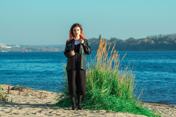 Portrait of beautiful stylish young woman with bright make-up, wearing black leather jacket and short culottes, standing on the river bank. 