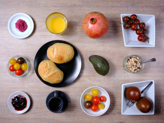 Pretzel, sweet tea-cake, marmelade, avocado, tomatoes, a pomegranate and a glass of orange juice, symetrically arranged on a wooden table on white and black tableware
