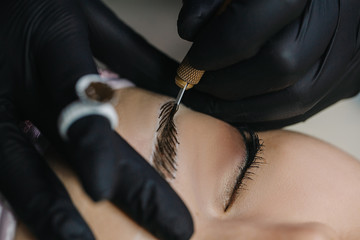 The master’s hand in a black glove holds a manipulation with a microblading needle over the model’s eyebrow, drawing hairs in macro photography.