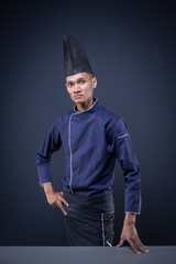 A portrait of an Asian executive chef in his blue uniform, black apron and black hat on dark grey background