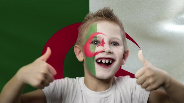Joyful fan on the background of the flag of Algeria. Happy boy with painted face in national colors. The young fan rejoices in the victory of his beloved team. Success. Victory. Triumph.