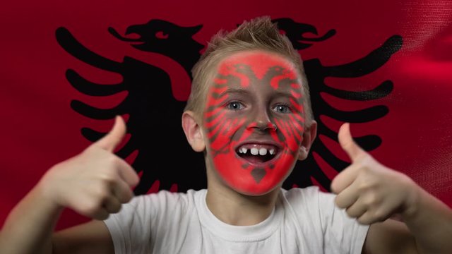 Joyful fan on the background of the flag of Albania. Happy boy with painted face in national colors. The young fan rejoices in the victory of his beloved team. Success. Victory. Triumph.