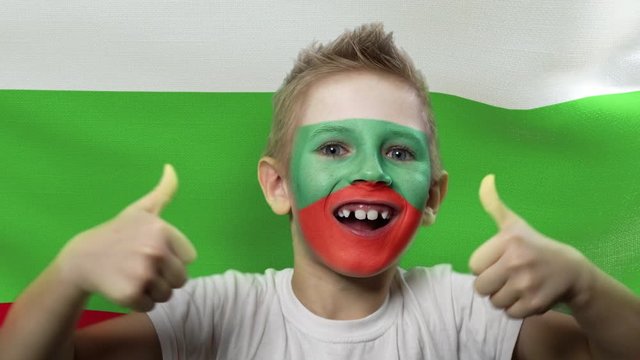 Joyful fan on the background of the flag of Bulgaria. Happy boy with painted face in national colors. The young fan rejoices in the victory of his beloved team. Success. Victory. Triumph.