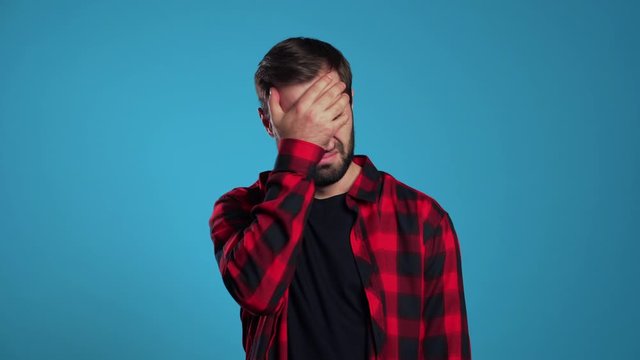 Young guy covers his face with hand from fatal disappointment, failure. Depressed lost man in red plaid shirt. Drama, emotions concept. 