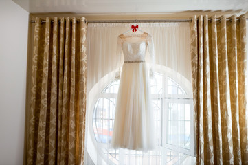 Amazing dress. Full length of beautiful silk wedding dress hanging near the window. Wedding day
