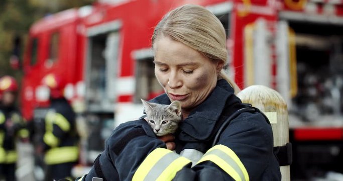 Portrait of the young blonde caucasian woman fire fighter holding and petting saved kitty cat from a fire. Outdoors.