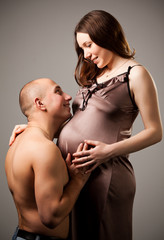 Man with naked torso hugging belly of pregnant woman dressed in nightie. Isolated on gray background