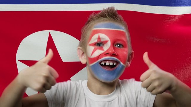 Joyful fan on the background of the flag of North Korea. Happy boy with painted face in national colors. The young fan rejoices in the victory of his beloved team. Success. Victory. Triumph.