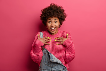 Excited surprised Afro American woman has happy reaction on received gift, keeps mouth opened, says is it for me, wears pink knitted sweater and denim dungarees, stands against bright studio wall