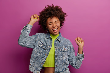 Triumphing happy curly woman celebrates finding new job, rasises arms in clenched fists, dressed in...