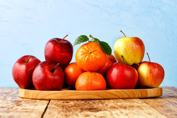 Red ripe apples and tangerines with green leaves lie on a wooden tray on an old wooden table with...