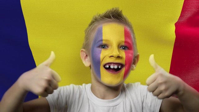 Joyful fan on the background of the flag of Romania. Happy boy with painted face in national colors. The young fan rejoices in the victory of his beloved team. Success. Victory. Triumph.
