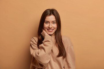 Confident woman has healthy skin, gentle smile, touches neck, expresses sincere emotions and happiness, feels encouraged, wears loose sweatshirt, isolated over brown background, looks friendly