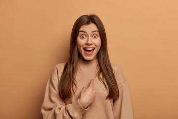Who, me? Portrait of beautiful surprised woman points at herself and looks with happy amazed expression, being picked or chosen, has long straight hair, wears casual sweatshirt, isolated on beige wall - obrazy, fototapety, plakaty