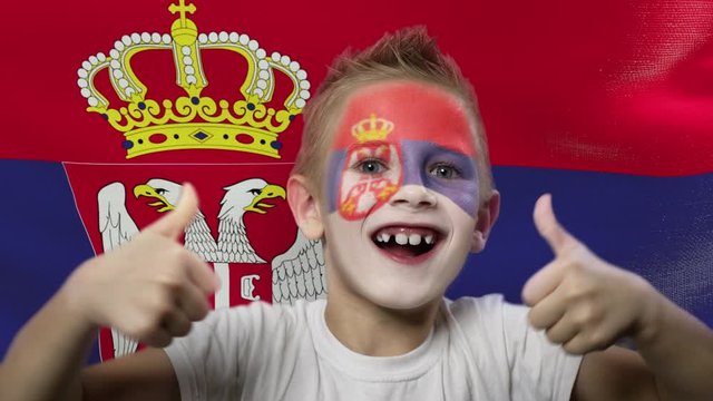 Joyful fan on the background of the flag of Serbia. Happy boy with painted face in national colors. The young fan rejoices in the victory of his beloved team. Success. Victory. Triumph.