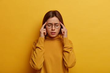 Unhealthy weak woman feels bad, suffers from strong migraine, keeps index fingers on temples, thinks abot something, tries to find solution, closes eyes, feels pressure, isolated on yellow background