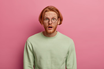 Stunned male with red hair and thick beard, keeps mouth opened, hears shocking rumors about friend, cant believe own eyes, holds breath from amazing news, wears spectacles and casual jumper.