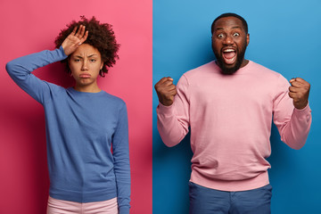 Tired African American woman feels overworked, rubs forehead, overjoyed bearded man clenches fists,...