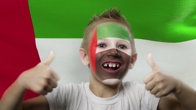 Joyful fan on the background of the flag of the UAE. Happy boy with painted face in national colors. The young fan rejoices in the victory of his beloved team. Success. Victory. Triumph.