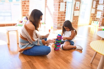 Beautiful teacher and toddler playing with wooden building blocks around lots of toys at kindergarten