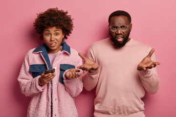 Displeased annoyed hesitant Afro American woman and man shrug shoulders with confused expressions, use mobile phone for online communication, express negative emotions, dressed in pink clothes