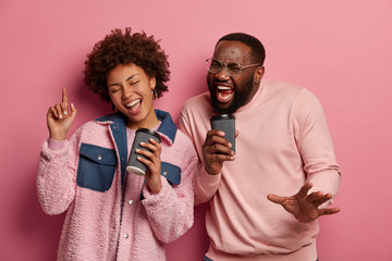 Photo of overjoyed dark skinned couple dance together, hold disposable paper coffee cups as microphones, sing songs, enjoys beverage dressed in casual wear, isolated on pink background, close eyes