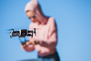 Standing with a bended knee, young woman in pink hoodie and dark glasses controls the drone's flight control panel.