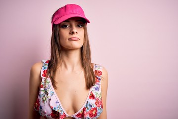Young beautiful brunette woman on vacation wearing bikini and cap over pink background with serious expression on face. Simple and natural looking at the camera.