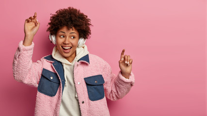 Emotional beautiful carefree teenage girl keeps hands up in air, dances with rhythm of music, wears wireless headphones, wears hoodie and jacket, looks somewhere, moves over rosy background.