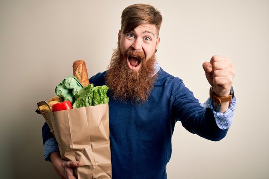 Redhead Irish man with beard holding fresh groceries from supermarkt over isolated background screaming proud and celebrating victory and success very excited, cheering emotion