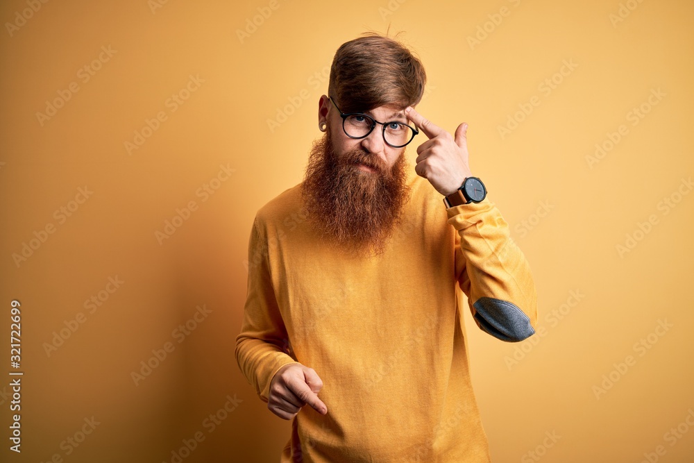 Poster Handsome Irish redhead man with beard wearing glasses over yellow isolated background pointing unhappy to pimple on forehead, ugly infection of blackhead. Acne and skin problem