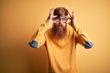 Handsome Irish redhead man with beard wearing glasses over yellow isolated background Trying to...