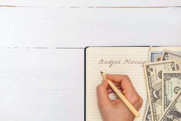 Budget planning concept. Directly above view of notepad with woman's hand writing words Budget Planning and some banknotes on white wooden background. Flat lay, copy space