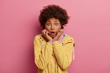 Fototapeta na wymiar Horizontal shot of shocked amazed Afro American woman keeps mouth opened, gasps with fear, notices something strange and very scary, wears yellow windbreaker, isolated over rosy studio background