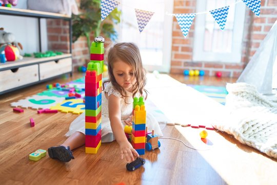 Adorable blonde toddler playing with building blocks around lots of toys at kindergarten