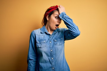 Young beautiful brunette woman wearing casual denim shirt and hair handkerchief surprised with hand...
