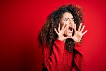 Young beautiful woman with curly hair and piercing wearing casual red sweater Shouting angry out loud with hands over mouth