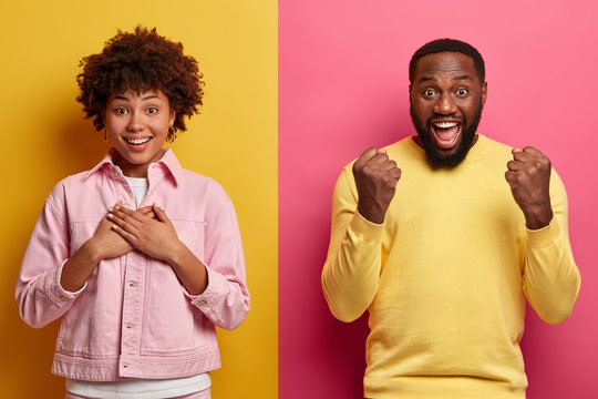 Portrait of pleased thankful woman keeps hands on chest, expresses gratitude and smile pleasantly, overjoyed bearded man clenches fists with triumph, express positive emotions. Afro American couple