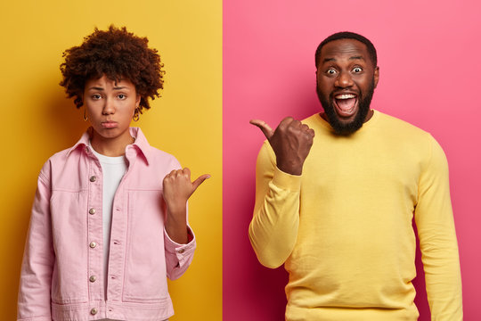 Offended displeased ethnic woman and happy bearded man point thumbs at each other, express their choice, dressed casually, expresses different emotions, isolated over rosy and yellow background
