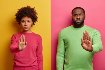 Serious looking dark skinned woman and man extend palms at camera, make stop warning gesture,...